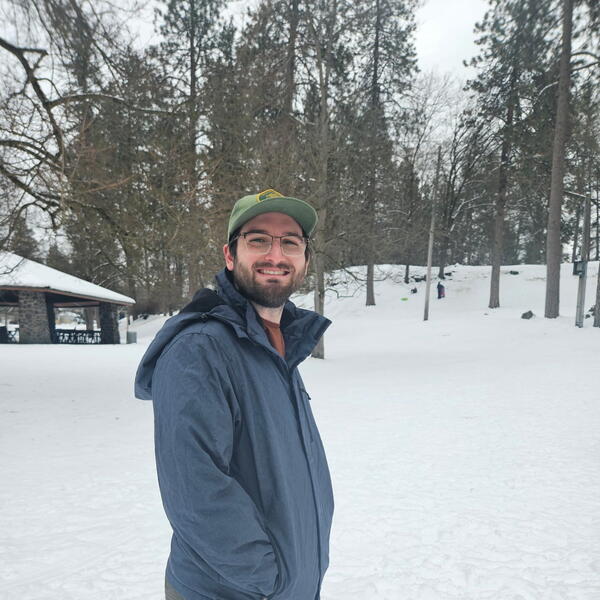 The image shows a man standing outdoors in a snowy landscape. He is smiling and looking at the camera. He is wearing a green baseball cap, glasses, a blue winter jacket, and olive green pants. His hands are in his jacket pockets. Behind him, there are tall pine trees, a snow-covered hill with a few people in the distance, and a covered picnic structure made of stone and wood. The atmosphere appears cold and wintry, with overcast skies and snow blanketing the ground and trees.